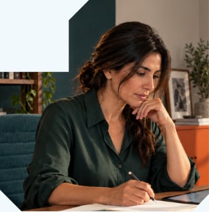 A woman writing in a notebook at her desk, chin in hand, deep in thought