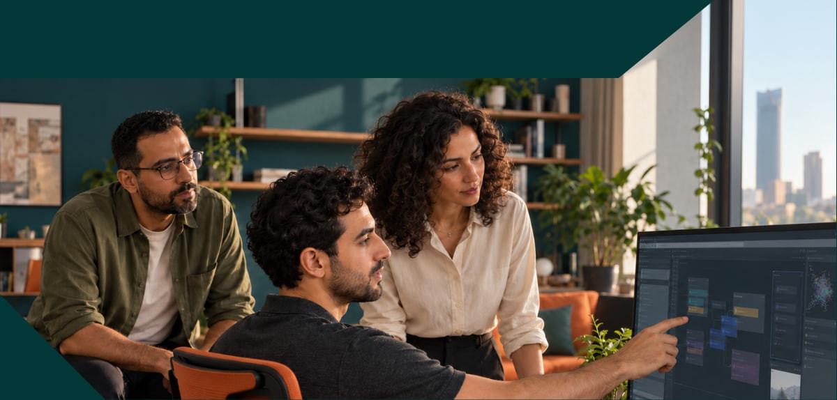 Three colleagues at a large monitor in an office, one pointing out a workflow on the screen